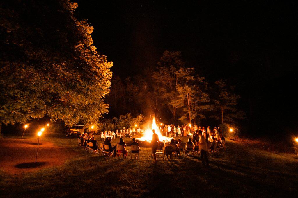 Nighttime outdoor gathering around a large bonfire with people sitting and standing, surrounded by trees.