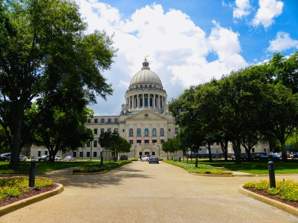 Capitol building with dome and surrounding trees in bright daylight.