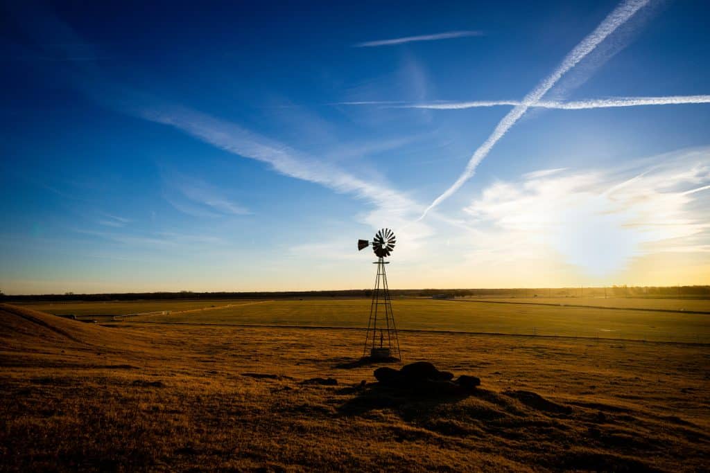 Sunset over open farmland featuring a windmill and expansive blue sky with contrails.