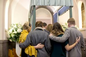 Group of family members standing together at a casket inside a chapel, arms around one another in a moment of support and reflection, representing church of christ funeral traditions.